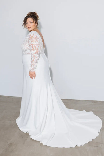 A woman in a white, embroidered florals lace-sleeved Julian - Jenny Yoo Wedding Dress with a train stands against a plain background, looking over her shoulder from Bergamot Bridal.