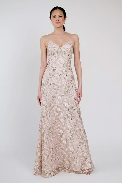 A woman is wearing the Julianna Bridesmaid Dress by Jenny Yoo from Bergamot Bridal, featuring a spaghetti strap, floor-length design made of romantic floral-patterned embroidered tulle fabric with a fitted bodice and flared skirt, standing against a plain background.