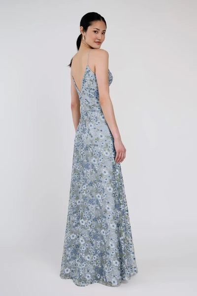 A woman in a blue floral Julianna Bridesmaid Dress by Jenny Yoo from Bergamot Bridal poses with her back slightly turned, showcasing the low-cut back and flowing design of the romantic style against a plain background.