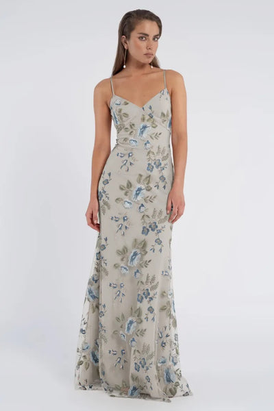 A woman wears a romantic, floor-length, floral-printed Julianna Bridesmaid Dress by Jenny Yoo from Bergamot Bridal with thin straps, posing against a plain white background.