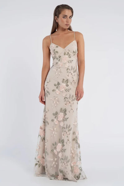 A woman stands wearing the romantic Julianna Bridesmaid Dress by Jenny Yoo from Bergamot Bridal—a floor-length, floral embroidered tulle gown with spaghetti straps. The light beige dress, adorned with pink and green detailing, creates an enchanting scene as she poses against a plain white background.