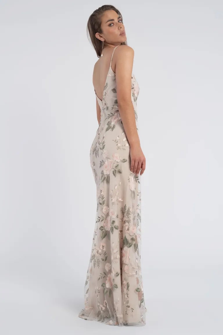 A woman in a romantic Julianna Bridesmaid Dress by Jenny Yoo from Bergamot Bridal stands sideways, looking over her shoulder. The long, sleeveless floral dress is light-colored with a pattern of pink flowers and green leaves, adding a touch of elegance to her look.