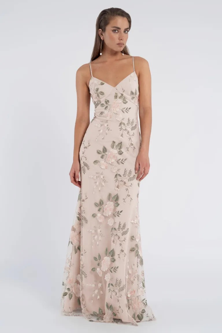 A woman stands wearing the Julianna Bridesmaid Dress by Jenny Yoo, a floor-length light pink gown with romantic floral embroidery, thin shoulder straps, and a V-neckline from Bergamot Bridal.