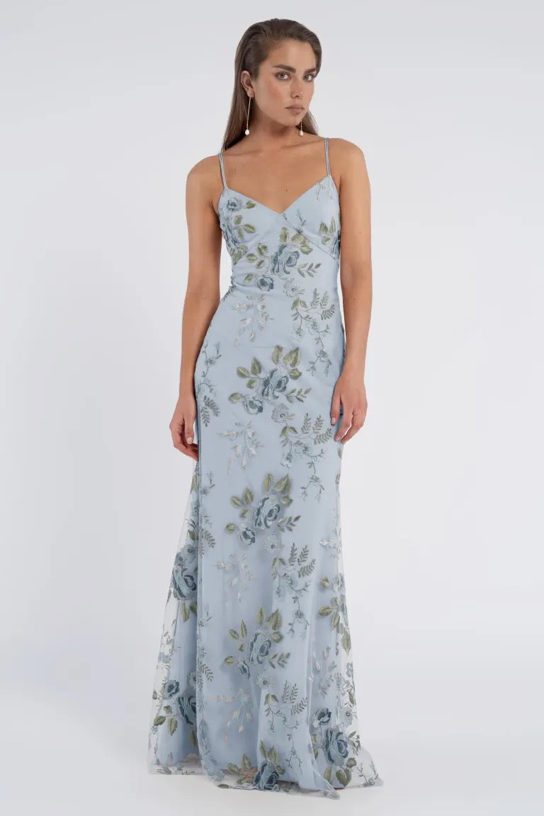 A woman stands wearing a long, light blue floral Julianna Bridesmaid Dress by Jenny Yoo from Bergamot Bridal, featuring thin straps, against a plain white background, exuding a romantic charm.