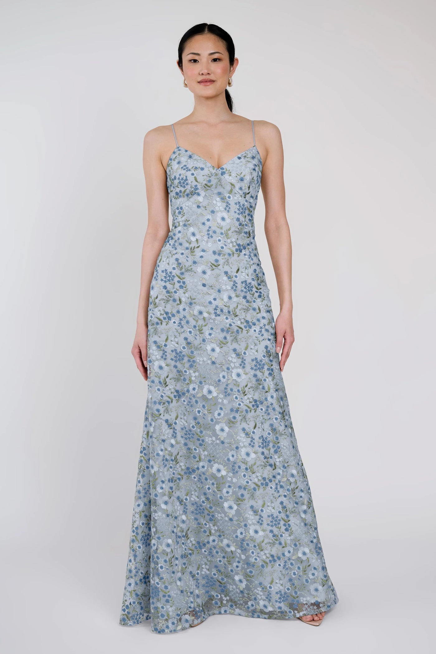 A woman stands wearing the Julianna Bridesmaid Dress by Jenny Yoo from Bergamot Bridal, a romantic long blue floral creation with spaghetti straps and delicate embroidered tulle fabric, set against a plain background.