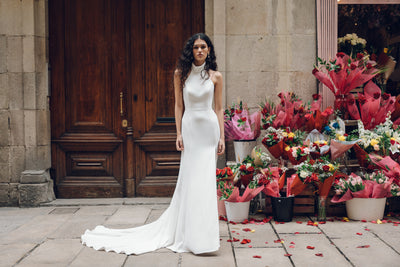 A woman wearing the Justine - Jenny Yoo Wedding Dress by Bergamot Bridal stands in front of a wooden door, accented by an arrangement of colorful flowers.