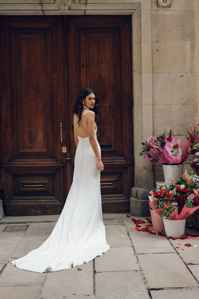 A woman wearing the Justine wedding dress by Bergamot Bridal, featuring an elegant fit and flare silhouette, stands in front of a wooden door with bouquets of flowers adorning the ground beside her.