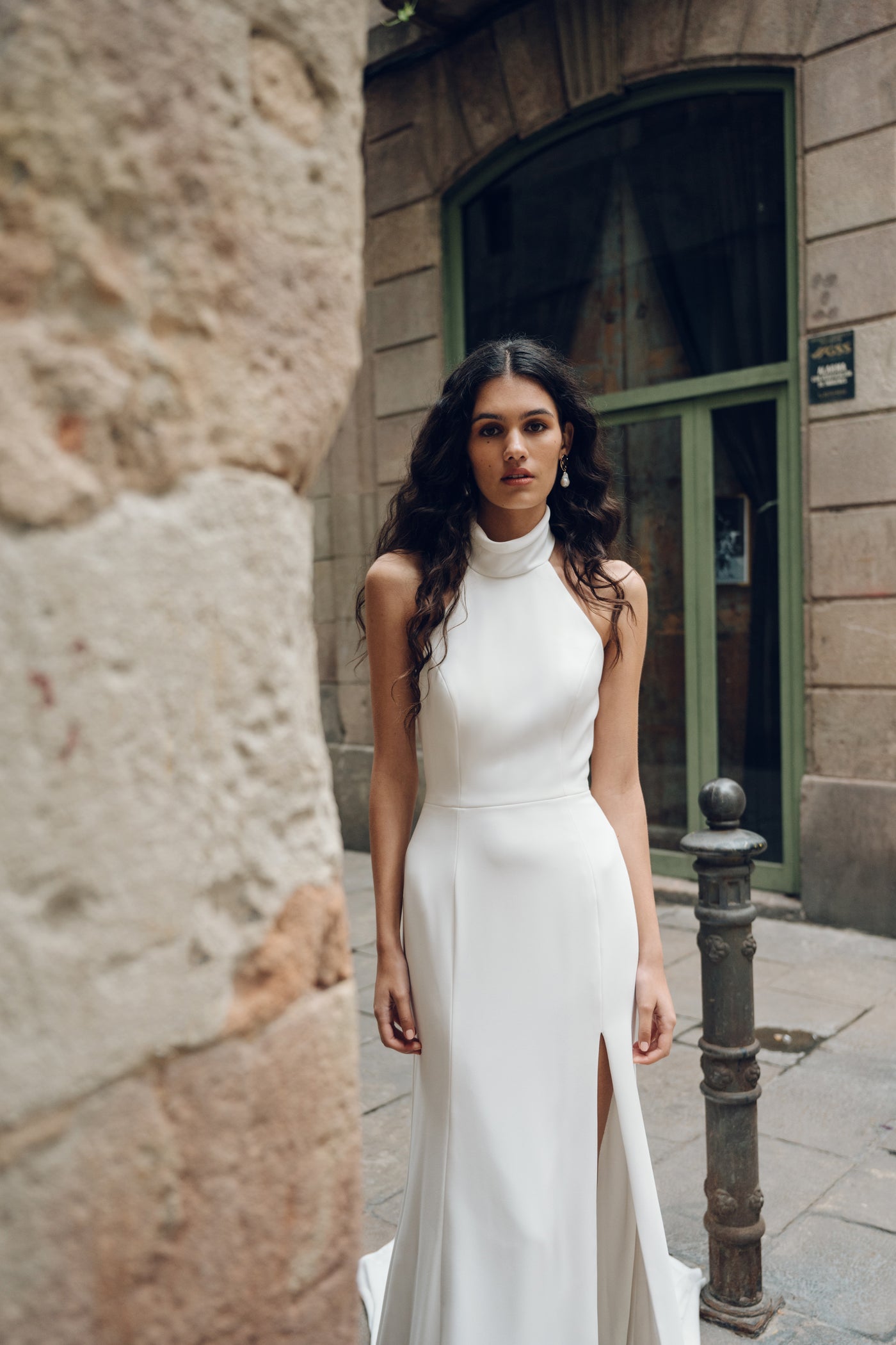 A woman in the Justine sleeveless wedding dress by Bergamot Bridal, which showcases a fit and flare silhouette with a high slit, stands in an urban alleyway beside a stone wall.