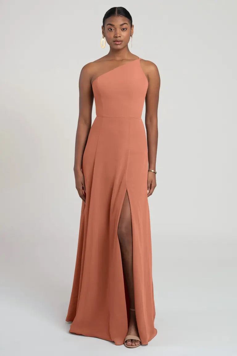 A woman in an elegant one-shoulder peach chiffon bridesmaid dress with a high slit, the Kora by Jenny Yoo from Bergamot Bridal.
