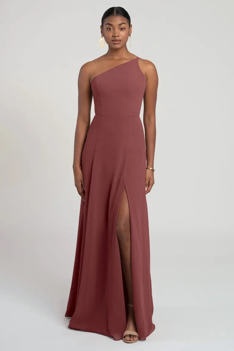 A woman posing in an elegant one-shoulder chiffon maroon evening gown with a side slit, the Kora - Jenny Yoo Bridesmaid Dress by Bergamot Bridal.