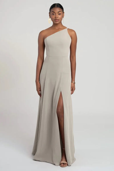 A woman poses in a flattering fit and flare silhouette one-shoulder chiffon bridesmaid dress with a high slit, the Kora - Jenny Yoo Bridesmaid Dress by Bergamot Bridal.