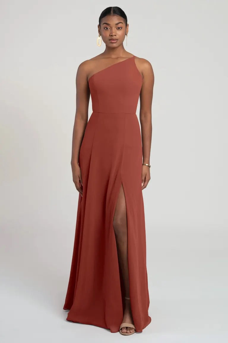 A woman wearing a one-shoulder, russet-colored chiffon Kora - Jenny Yoo bridesmaid dress with a high slit from Bergamot Bridal.