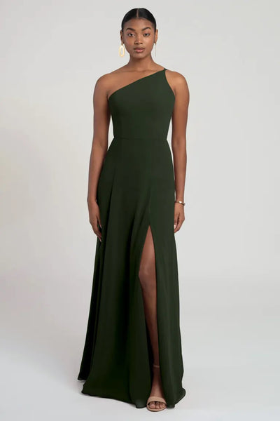 A woman stands wearing the Kora - Jenny Yoo Bridesmaid Dress from Bergamot Bridal, an asymmetrical dark green gown with a high slit. She accessorizes with gold earrings and an elegant bracelet, set against a plain background.