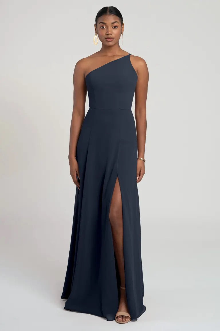 A woman standing against a neutral background, wearing a Kora - Jenny Yoo bridesmaid dress in navy blue with a slit, complemented by drop earrings and open-toed shoes from Bergamot Bridal.