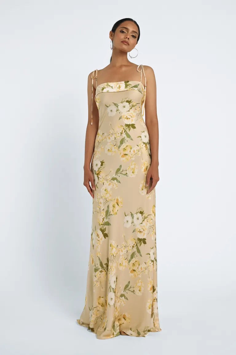 A woman is standing against a white background, wearing the Landry Bridesmaid Dress by Jenny Yoo from Bergamot Bridal, a long, sleeveless floral dress with spaghetti straps and a fitted bodice.