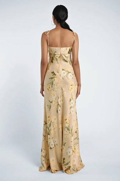 A woman is standing with her back facing the camera, wearing the Landry Bridesmaid Dress by Jenny Yoo featuring a long, floral pattern with sleek neckline bands and shoulder ties, from Bergamot Bridal.