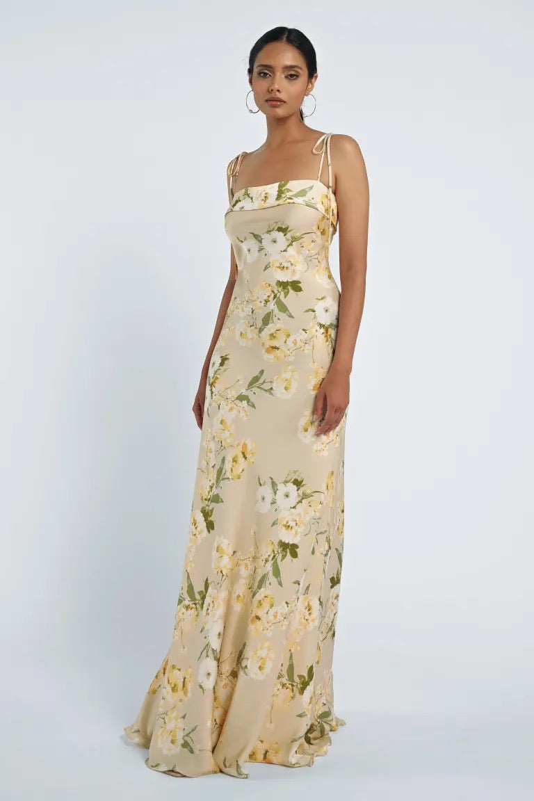 A person is wearing the Landry Bridesmaid Dress by Jenny Yoo from Bergamot Bridal, a long, cream-colored floral dress with a sleek neckline band and spaghetti straps. They are standing against a plain white background.