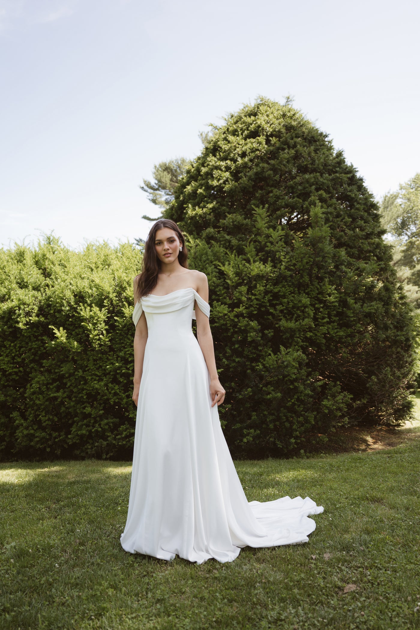 A woman in a Lee by Jenny Yoo gown from Bergamot Bridal with detachable swag sleeves stands on grass before large green bushes in the sun.