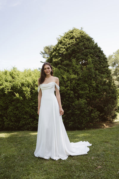 A woman in a Lee by Jenny Yoo gown from Bergamot Bridal with detachable swag sleeves stands on grass before large green bushes in the sun.
