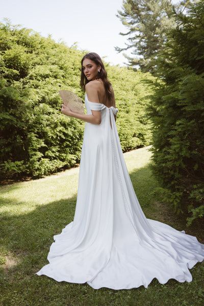 Wearing the Lee by Jenny Yoo gown from Bergamot Bridal, a woman stands on grass holding a fan, her classic silhouette with detachable swag sleeves framed by tall green hedges.