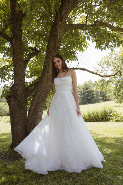 A woman in the Lorelai by Jenny Yoo gown from Bergamot Bridal stands outdoors on grass near a tree, sunlight filtering through the leaves.