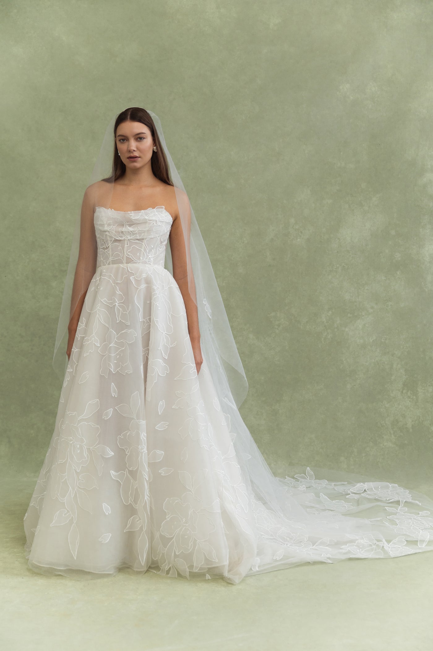 A woman stands before a green backdrop in a strapless white wedding dress with oversized floral appliqué, wearing the Lorelai Veil by Jenny Yoo from Bergamot Bridal trailing elegantly behind her.
