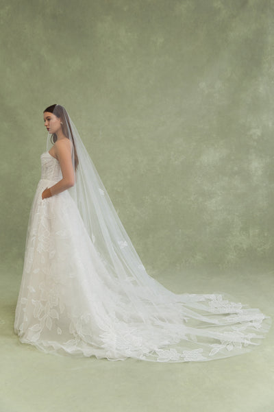 A woman in a strapless white wedding dress with oversized floral appliqué and a long, sheer Lorelai Veil by Jenny Yoo from Bergamot Bridal stands against a muted green background.