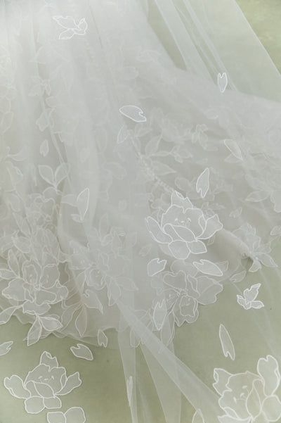 The Lorelai Veil by Jenny Yoo from Bergamot Bridal features sheer white fabric embroidered with large floral blossoms and leaves, evoking a romantic wedding veil, displayed on a light-colored surface.