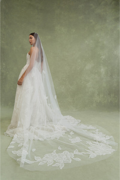 A woman in a strapless white wedding gown with an oversized floral applique and the Lorelai Veil by Jenny Yoo from Bergamot Bridal stands against a green textured background, facing slightly away from the camera.