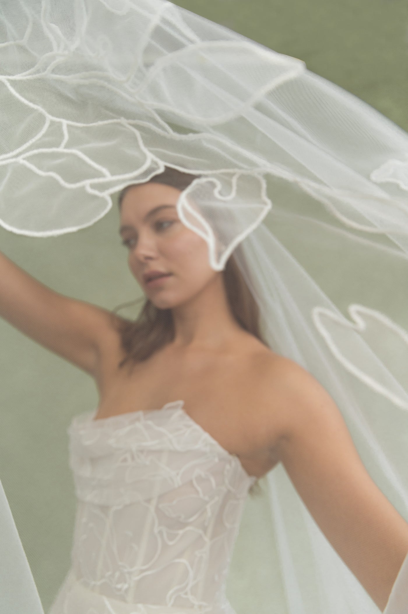A woman wearing a white, textured strapless dress and the Lorelai Veil by Jenny Yoo from Bergamot Bridal stands against a muted green background.