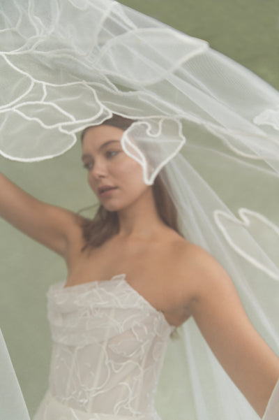 A woman wearing a white, textured strapless dress and the Lorelai Veil by Jenny Yoo from Bergamot Bridal stands against a muted green background.