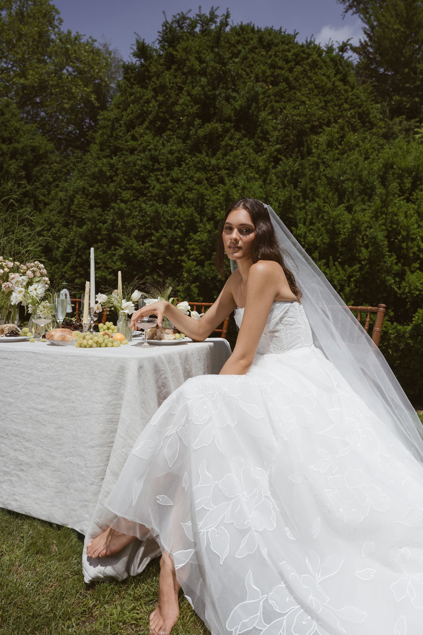 A bride in the Lorelai by Jenny Yoo gown from Bergamot Bridal sits barefoot at an outdoor table adorned with flowers, candles, and food, greenery in the background.