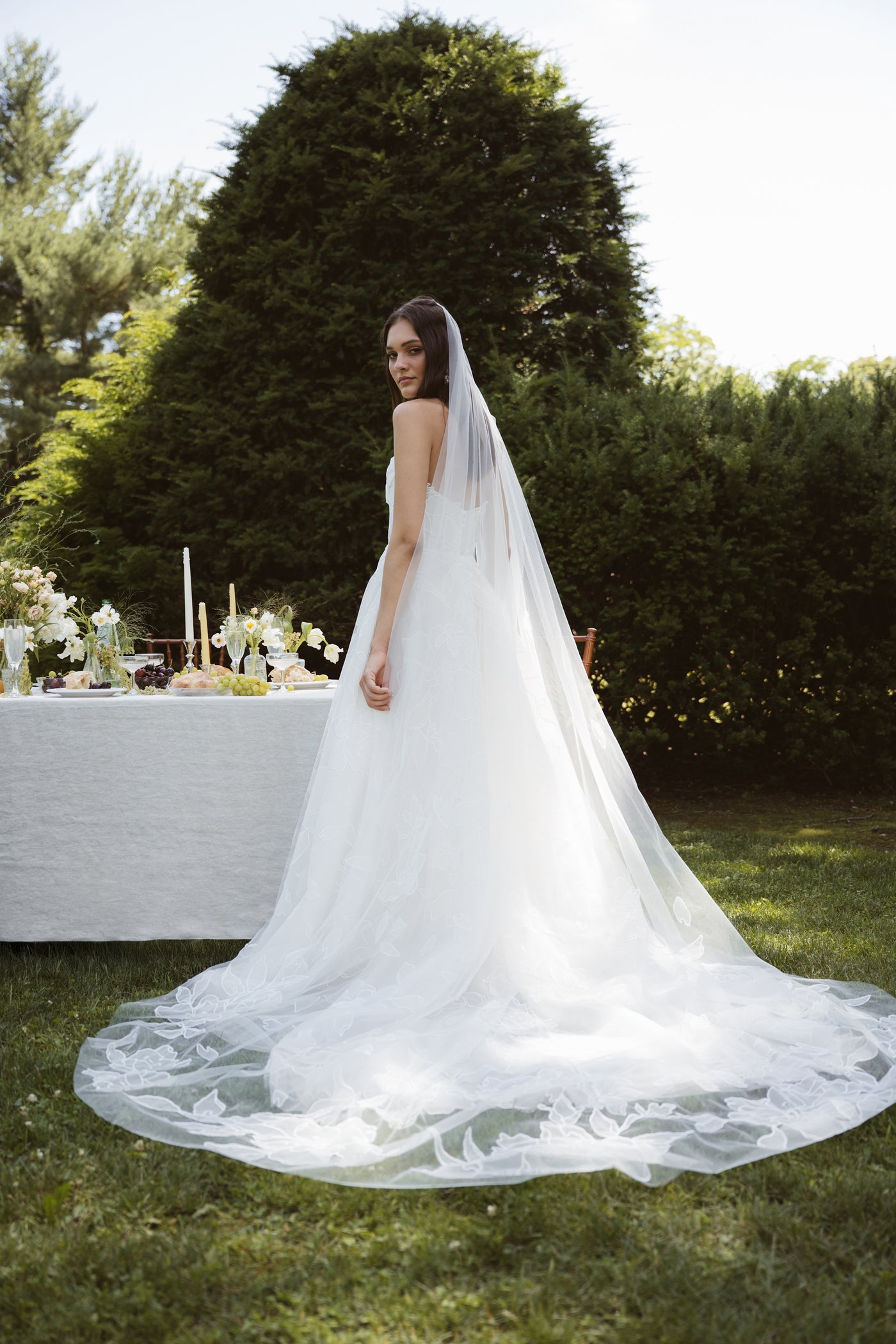 A woman in the Lorelai by Jenny Yoo gown from Bergamot Bridal, featuring delicate floral appliques and a long veil, stands on grass near a decorated outdoor table with greenery and trees in the background.
