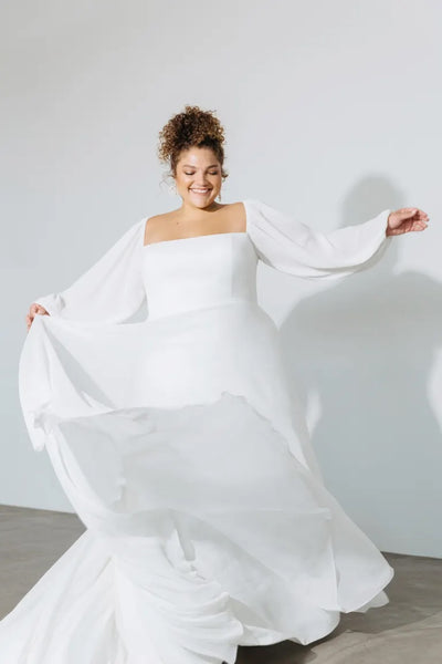 A woman in a flowing white wedding dress with a chiffon fabric skirt smiling and twirling the Louise - Jenny Yoo Wedding Dress by Bergamot Bridal.