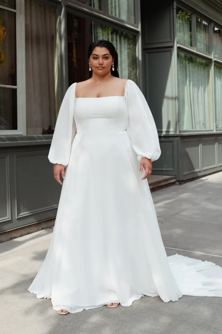 A woman in a white Louise - Jenny Yoo wedding dress with an A-line skirt and long sleeves standing in front of a building.
