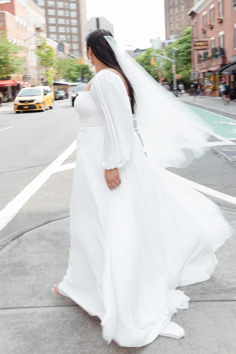 A bride in a Louise - Jenny Yoo wedding dress and long veil walking on a city street.