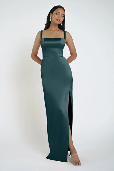 A woman stands against a plain background wearing a dark green satin Lowe Bridesmaid Dress by Jenny Yoo from Bergamot Bridal, featuring a thigh-high slit and square neckline. She accessorizes with minimal jewelry, including drop earrings.