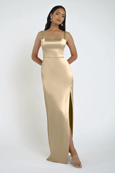 A woman in a sleeveless, floor-length, champagne-colored Lowe Bridesmaid Dress by Jenny Yoo from Bergamot Bridal stands against a plain white background. She has long hair, is wearing earrings and heels, and the dress features a square neckline that adds an elegant touch.