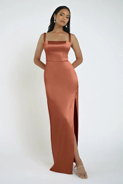 A woman stands in the Lowe Bridesmaid Dress by Jenny Yoo, a sleeveless, floor-length rust-colored gown featuring a high slit and square neckline, posing against a plain white background. The dress is from Bergamot Bridal.