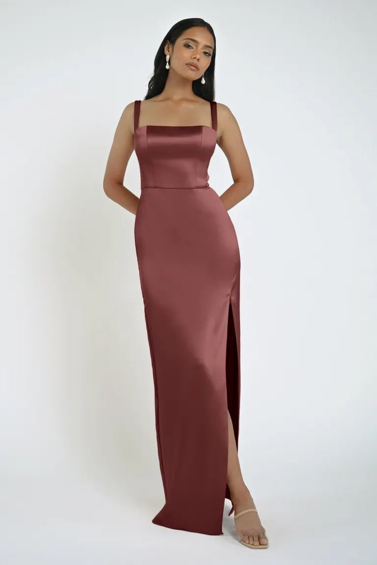 A woman stands wearing the Lowe Bridesmaid Dress by Jenny Yoo, a sleeveless burgundy satin gown with a square neckline and a sleek skirt featuring a thigh-high slit, paired with nude heels.