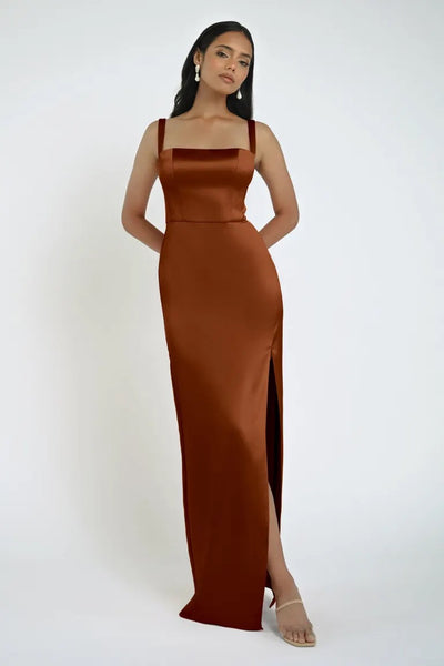 A woman stands with hands behind her back, wearing a sleeveless, copper-colored Lowe Bridesmaid Dress by Jenny Yoo from Bergamot Bridal, featuring a sleek skirt and slit. She has long dark hair and is accessorized with earrings and open-toe heels.