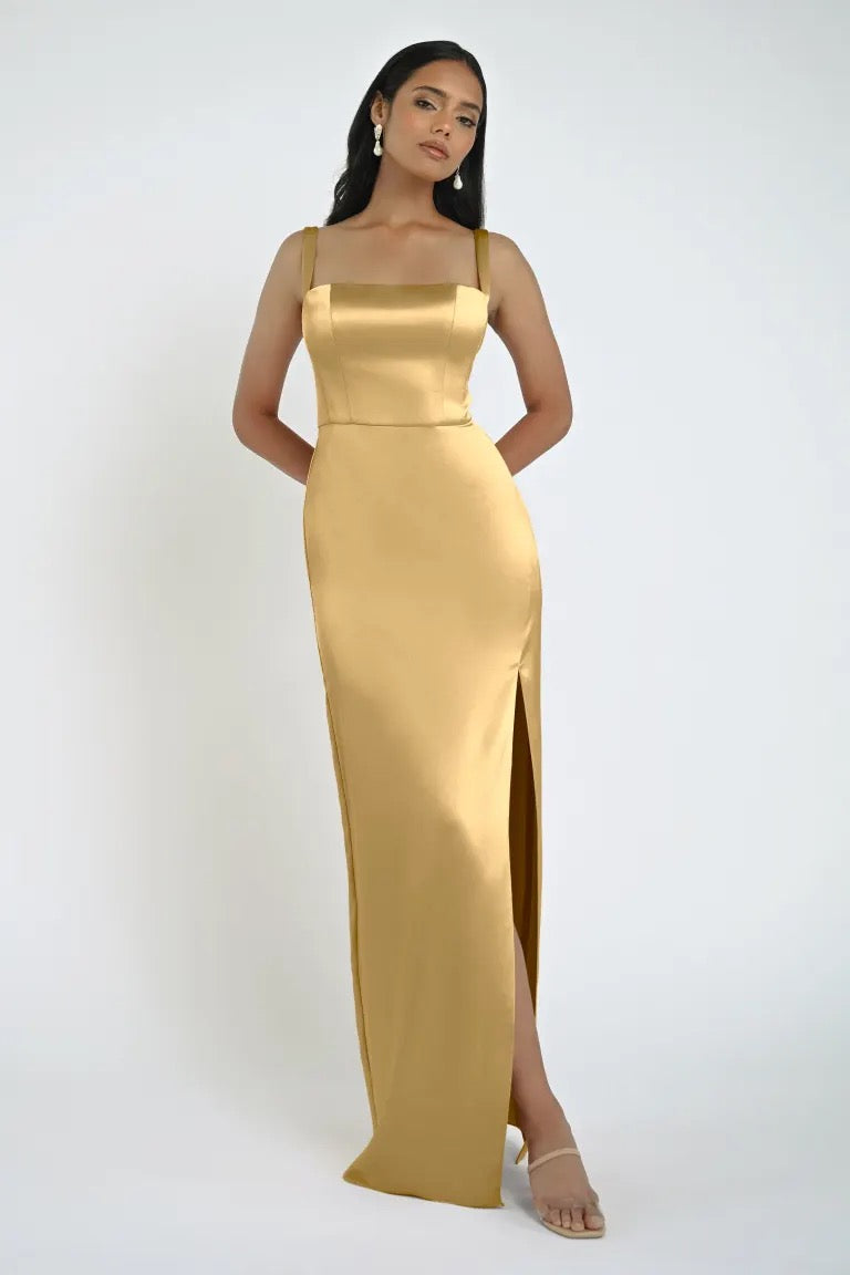 A woman wearing a sleeveless Lowe Bridesmaid Dress by Jenny Yoo in a gold hue, featuring a sleek skirt and square neckline, poses against a plain background.