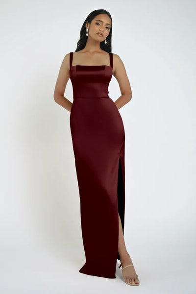 A woman is standing in a burgundy Lowe Bridesmaid Dress by Jenny Yoo from Bergamot Bridal, featuring a square neckline and a high slit on one side. She pairs the elegant dress with nude heels and minimal jewelry against a plain white background.