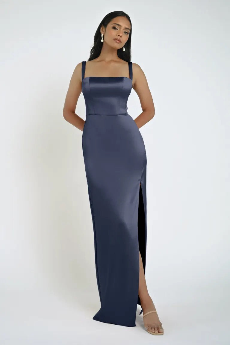 A woman stands against a plain background wearing a fitted, floor-length Lowe Bridesmaid Dress by Jenny Yoo with a sleek skirt and square neckline. She has her hands behind her back and is wearing earrings and open-toe heels from Bergamot Bridal.