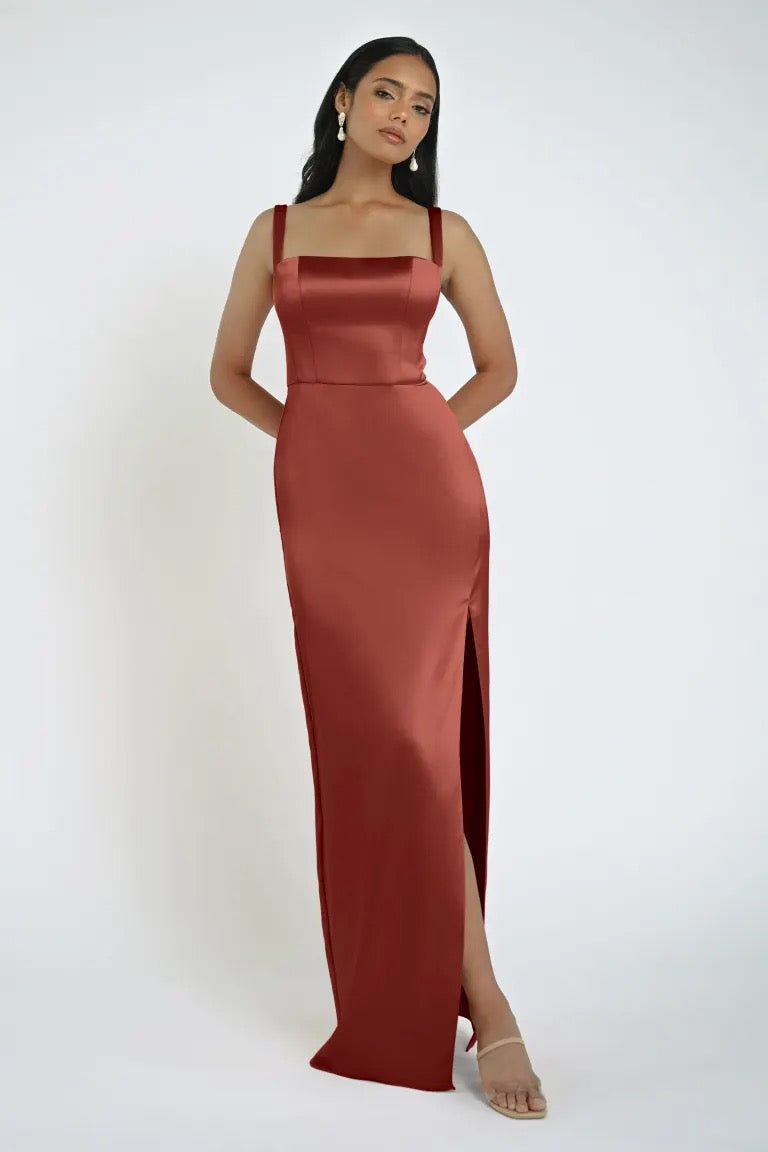 A woman stands in a Bergamot Bridal Lowe Bridesmaid Dress by Jenny Yoo—a red satin evening gown with a high slit and square neckline—posing against a plain white background.