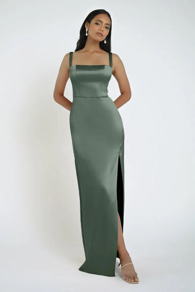 A woman in a sleeveless dark green satin Lowe Bridesmaid Dress by Jenny Yoo from Bergamot Bridal poses against a white background. The dress features a square neckline, fitted bodice, and a thigh-high slit. She is accessorized with earrings and open-toe heels.