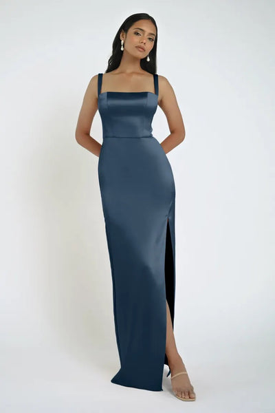 A woman stands in a blue, sleeveless, floor-length Lowe Bridesmaid Dress by Jenny Yoo from Bergamot Bridal, featuring a daring slit. She is wearing dangling earrings and open-toe heels.