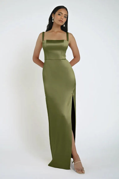 A woman wearing an olive green Lowe Bridesmaid Dress by Jenny Yoo from Bergamot Bridal, featuring a daring slit and square neckline, poses against a light gray background. She has long, dark hair, is adorned with earrings, and sports clear high-heeled shoes.