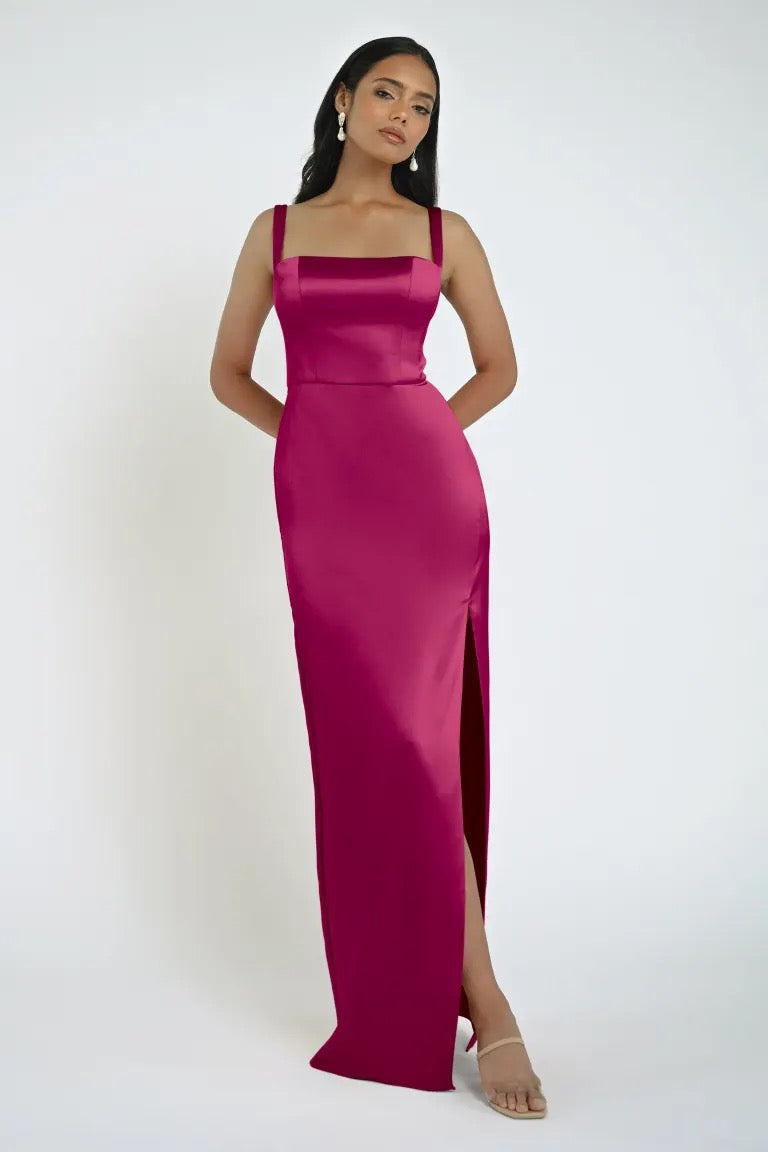 Woman wearing the Lowe Bridesmaid Dress by Jenny Yoo from Bergamot Bridal, a fitted, magenta floor-length gown with a sleek skirt and square neckline, posing with one leg forward and hands relaxed at her sides in front of a white background.