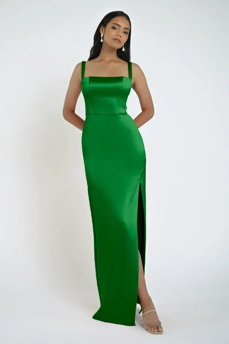 A woman in a bright green, floor-length Lowe Bridesmaid Dress by Jenny Yoo, sold by Bergamot Bridal, stands against a plain white background. She has long black hair and is wearing earrings. The dress features a high slit and square neckline.